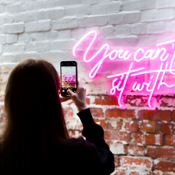 Person taking a photo of a pink LED neon sign reading You Can't Sit with Us - Custom Neon