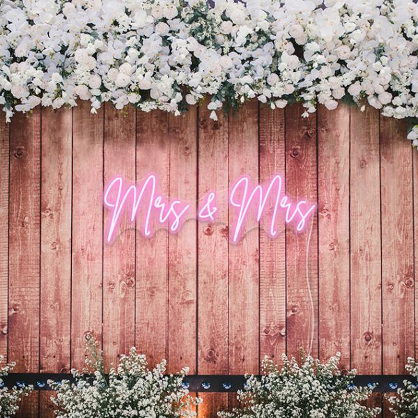 Mrs & Mrs CUSTOM NEON® LGBTQ+ Wedding Sign in soft pink on rustic wooden background surrounded by white & pink flowers