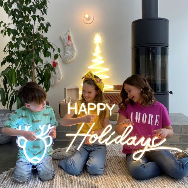 CUSTOM NEON® Holiday Lights: Reindeer Face, Happy Holidays & Modern LED Neon Xmas Tree held by children in front of a Nativity scene, stockings & wood burning stove