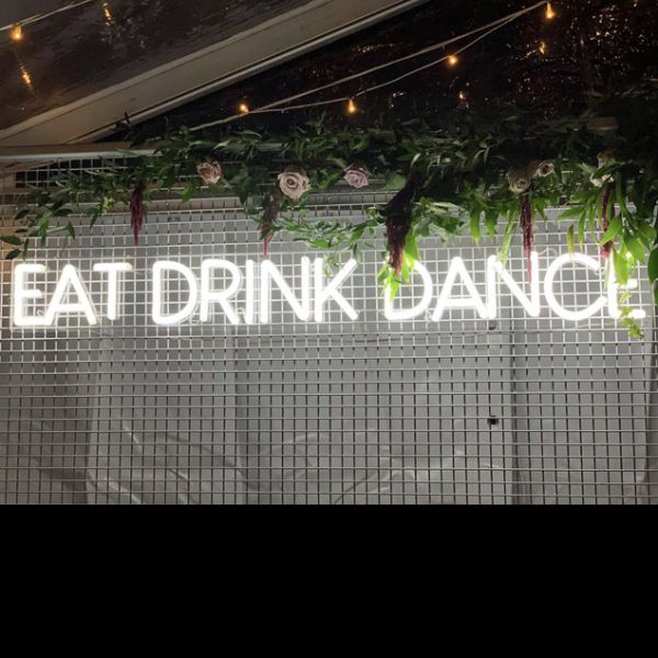 EAT DRINK DANCE large LED neon sign for events & weddings, shown on wire mesh with floral arrangement & fairy lights - photo from CustomNeon.com