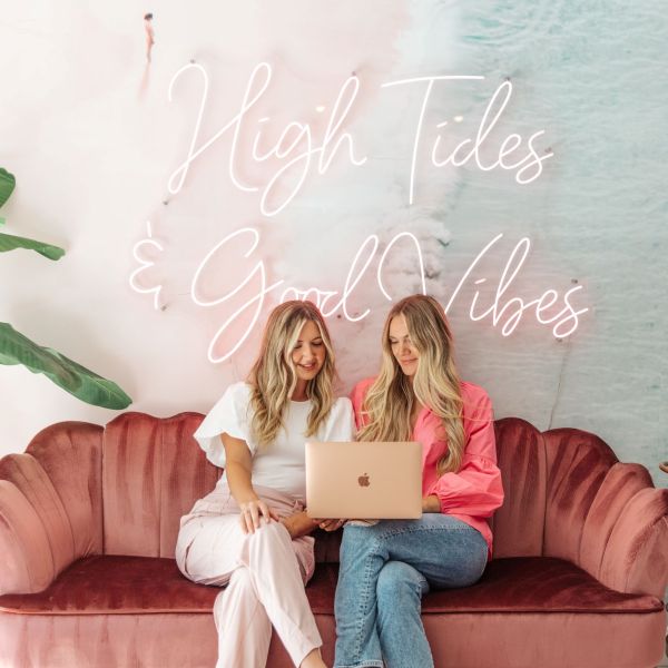 High Tides & Good Vibes pink LED neon sign installed a soft pink and blue wall mural of waves on the beach with two women looking at a laptop in the foreground - CUSTOM NEON™ x SOMERLED COLLECTION