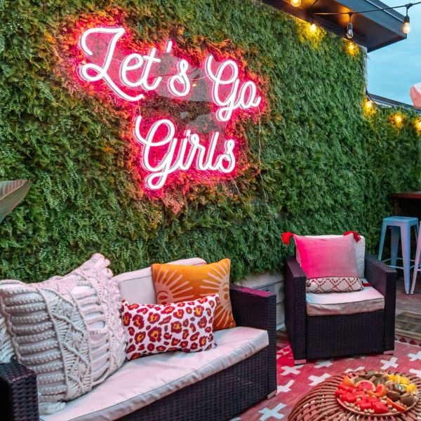 Let's Go Girls pink LED neon sign on a lush greenery wall in an outdoor seating area - CUSTOM NEON™ x SOMERLED COLLECTION