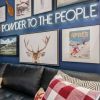 Powder to the People white LED neon sign on a blue feature wall surrounded by framed paintings and prints of mountain scenes - CUSTOM NEON™ x SOMERLED COLLECTION