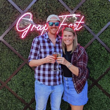 Couple with champagne in front of the pink GrapeFest script LED neon sign on a hedge wall in Grapevine, Texas

