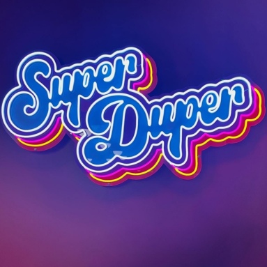Blue Super Duper script LED neon sign with a rainbow layered acrylic backing on a purple wall