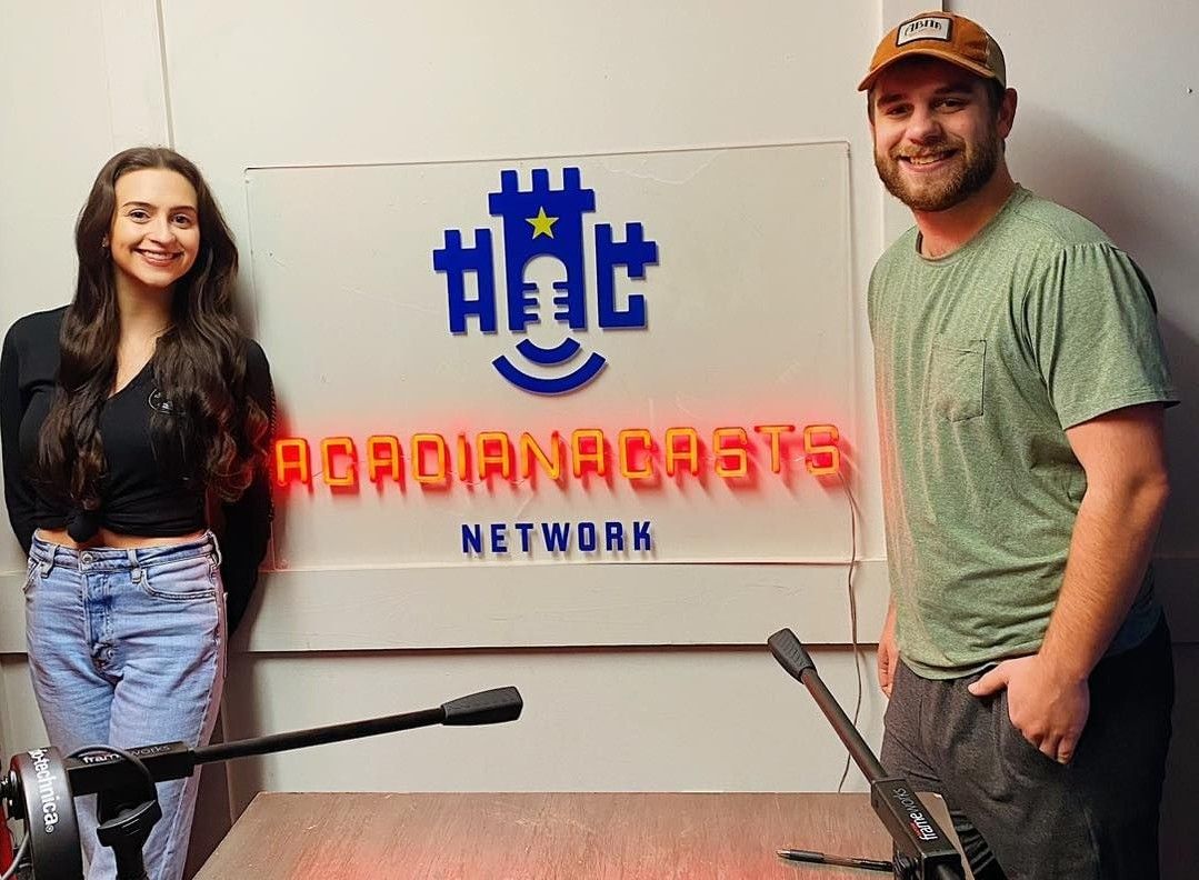 Man and woman standing on either side of a custom neon 'AcadianaCasts Network' sign with a castle and microphone logo in a podcast studio