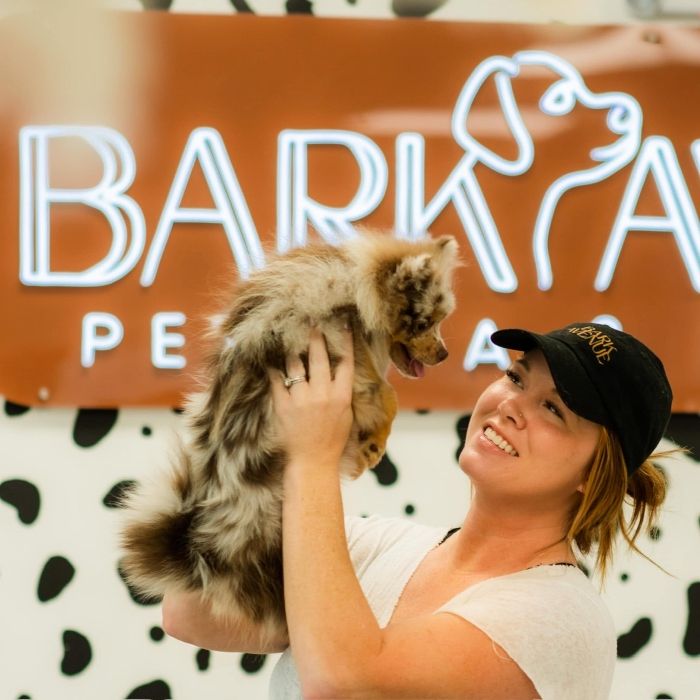 Woman holding up a fluffy Australian shepherd puppy in front of a Custom Neon® Bark Avenue Pet Spa sign