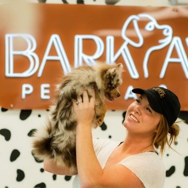 Woman holding up a fluffy Australian shepherd puppy in front of a Custom Neon® 'Bark Avenue Pet Spa' sign