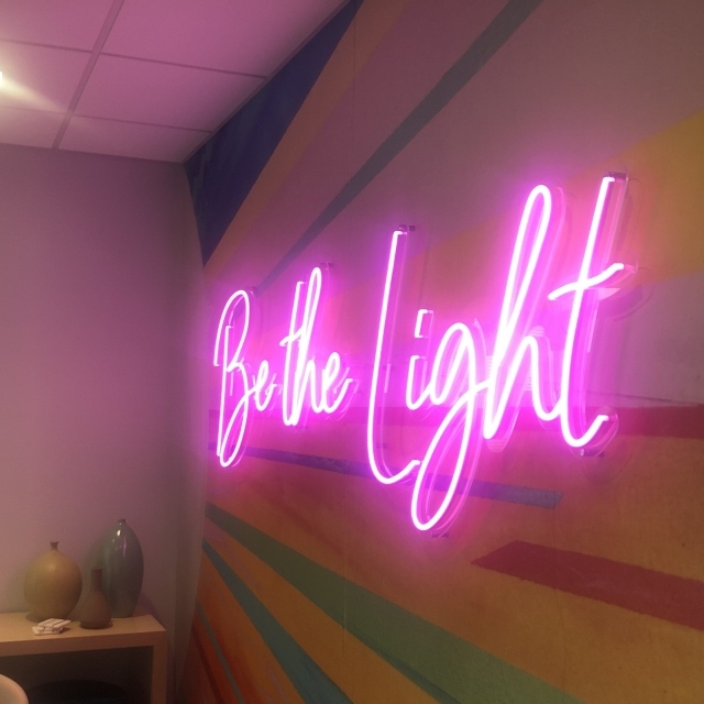Pink custom neon 'Be the Light' script sign on a wall with a colorful rainbow sunburst mural