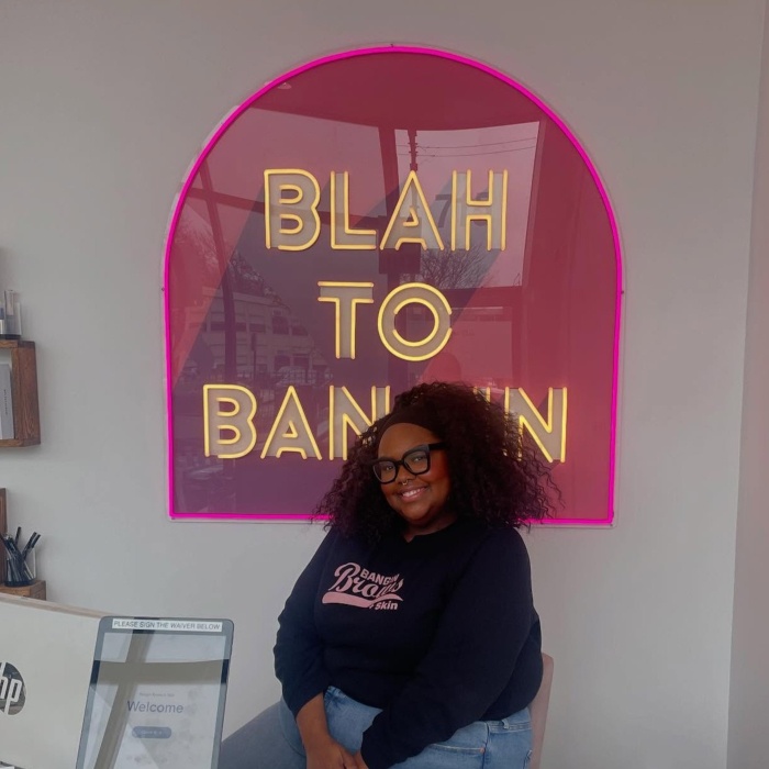 Blah to Bangin Custom Neon® sign in pink and gold above a smiling Bangin Brows client