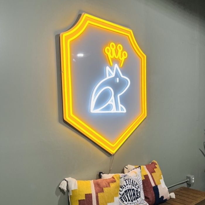 Custom logo LED neon sign of a crowned frog inside a yellow shield, mounted on a gray wall above cushions at Casa Rana