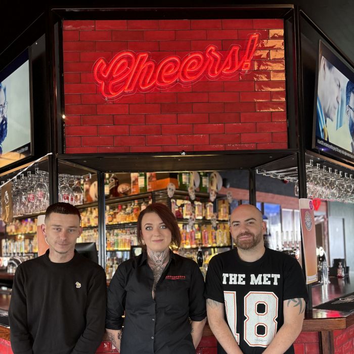 Three Davenports bar staff standing beneath a custom red LED neon 'Cheers!' sign on a red brick-tiled wall