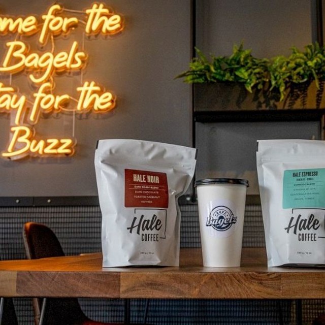 Yellow Custom Neon® sign on a black wall featuring the @buzzbagelz tagline