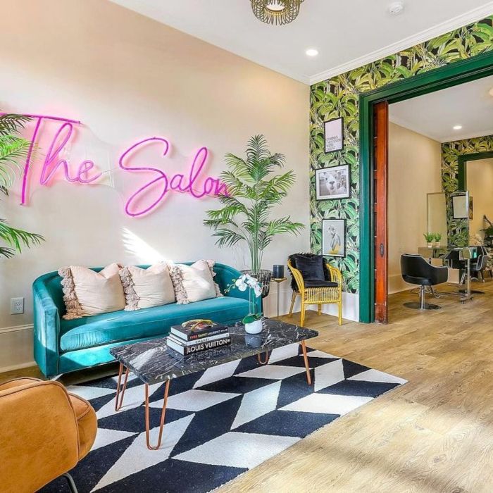 Pink Custom Neon® 'The Salon' script sign above a teal velvet sofa in a stylish salon lounge with tropical decor
