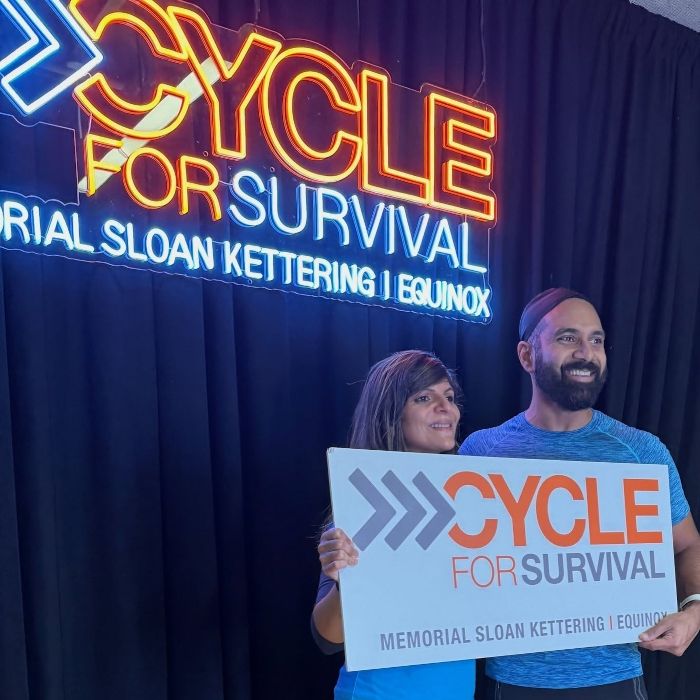 Couple holding a Cycle for Survival sign beneath the event's orange and blue LED neon backdrop for Memorial Sloan Kettering