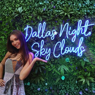 Woman posing next to a blue Custom Neon® Dallas Night Sky Clouds sign on a grass wall