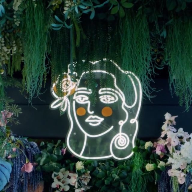 Delbar's white and orange LED neon lady logo glowing among hanging plants and flowers