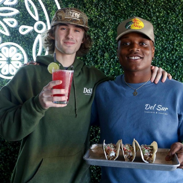 White Custom Neon® Mexican sugar skull logo on a grasswall with patrons posing for a selfie in the foreground @delsurtacqueria