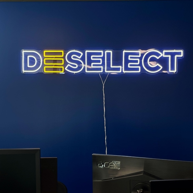 Custom Neon® 'Deselect' sign with yellow accent detail on a navy office wall above two computer monitors