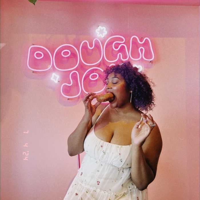 Dough Joy pink Custom Neon® sign in a double-lined custom font with a customer eating a donut in the foreground @doughjoydonuts