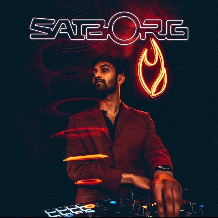 DJ performing under red light in front of an orange flame neon and a custom neon 'Satzorg' logo sign