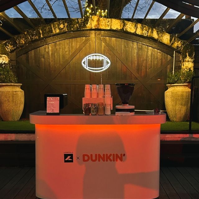 White football outline LED neon sign above a Learfield x Dunkin' coffee bar at an outdoor Dallas event
