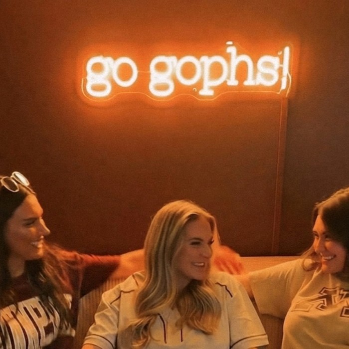 Yellow Custom Neon® sign reading Go Gophs on the wall in the background of women on a couch at The Gopher House