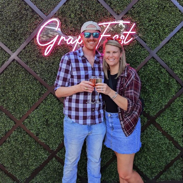 Couple with champagne in front of the pink GrapeFest script LED neon sign on a hedge wall in Grapevine, Texas
