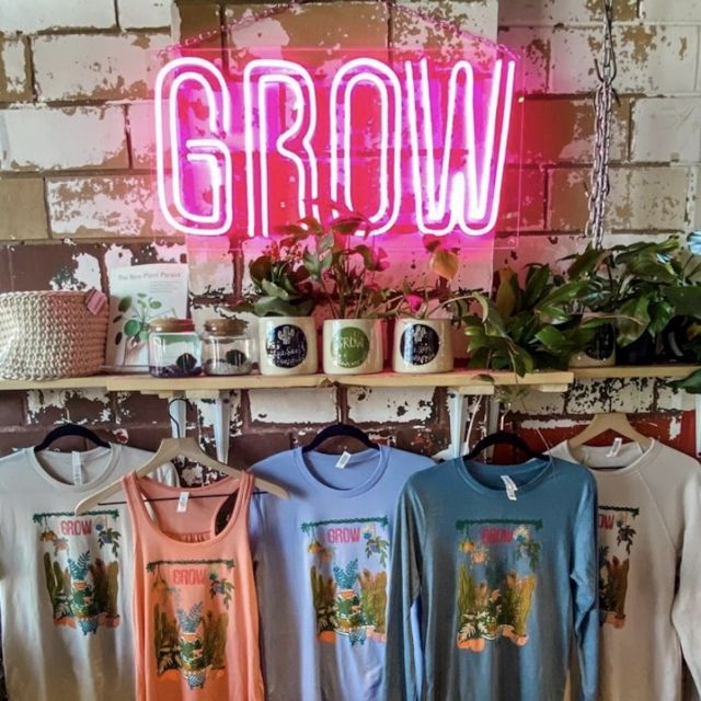 GROW Custom Neon® sign in pink LED neon and a double-lined font. Mounted on a distressed brick wall with plants and merchandise in the foreground growgiesenplantshop.com @botanicatgrow @growplantbar