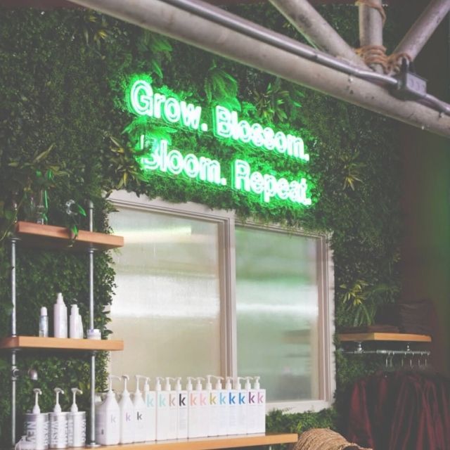 Grow Blossom Bloom Repeat green LED neon sign on a greenery wall above salon haircare shelves