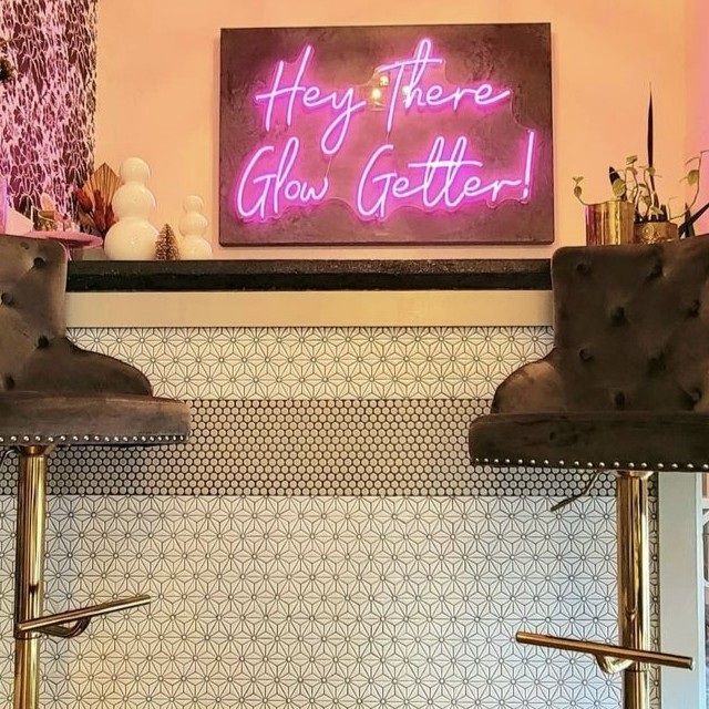 Pink Custom Neon® 'Hey There Glow Getter!' sign above a pair of black velvet bar stools in a salon with patterned tile
