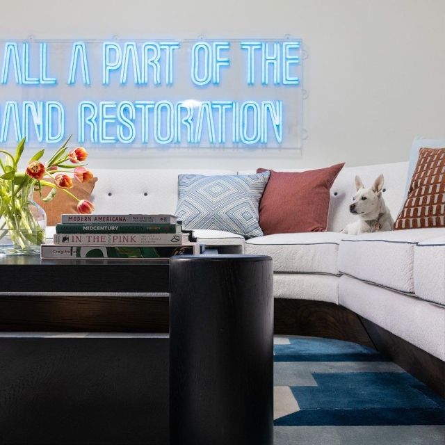 Blue LED neon quote sign reading It's All a Part of the Grand Restoration above a sofa with a dog