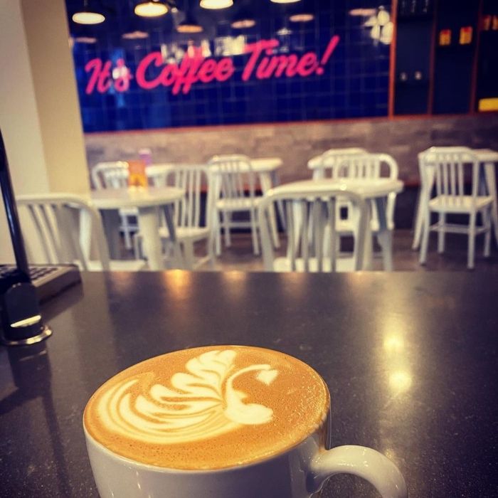 Latte with leaf latte art on a counter, with a pink custom neon 'It's Coffee Time!' sign on a blue tiled wall behind
