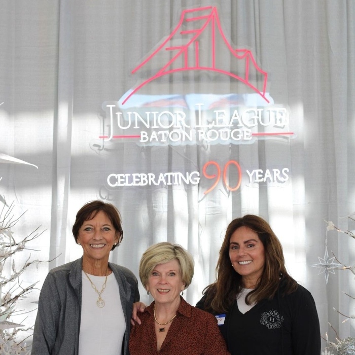 Three women in front of a custom neon 'Junior League Baton Rouge, Celebrating 90 Years' sign on a white curtain backdrop