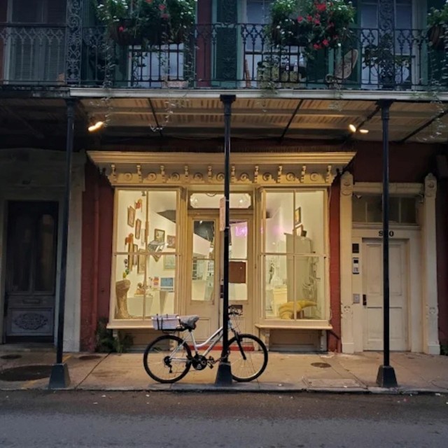 Evening street view of a French Quarter gallery storefront with lit display windows, a pink neon sign glowing inside, and a bicycle leaning against a lamppost