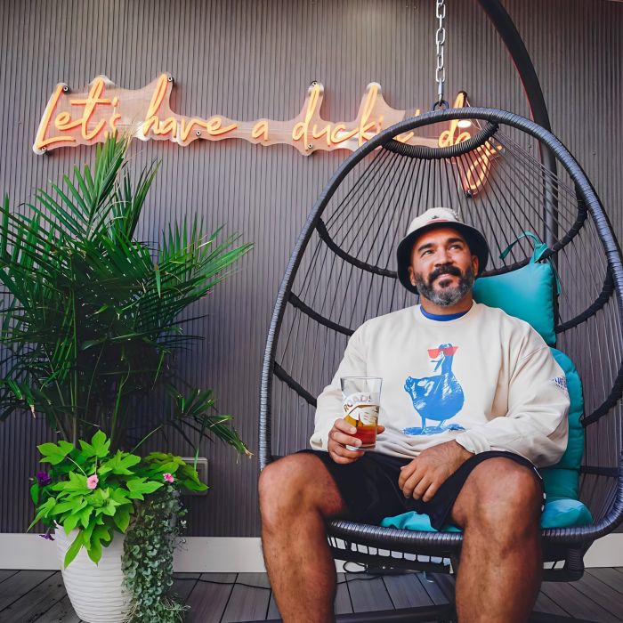 Man sitting on a swing chair holding a drink in front of an orange an orange Lets Have a Duckin Day neon sign at The Blue Duck in New London CT