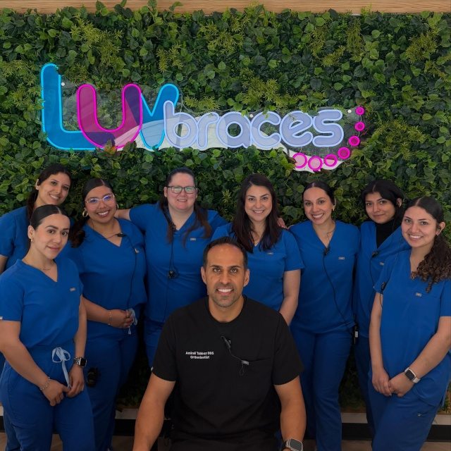 LUVbraces orthodontic team in blue scrubs with their orthodontist, in front of a green foliage wall and custom 'LUVbraces' logo sign