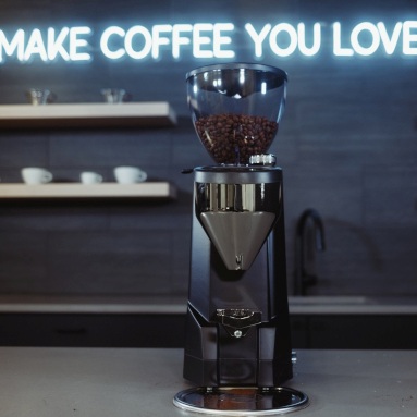 Make Coffee You Love white Custom Neon® slogan sign on a black wall  with a coffee grinder in the foreground @seattlecoffeegear