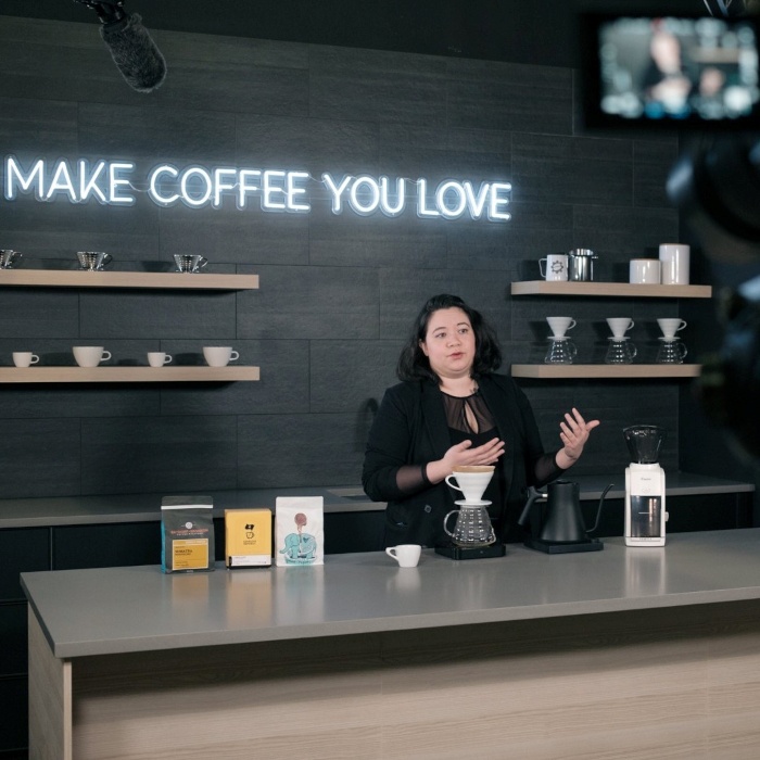 Make Coffee You Love white Custom Neon® quote sign on a black wall with a person talking into a video camera @seattlecoffeegear