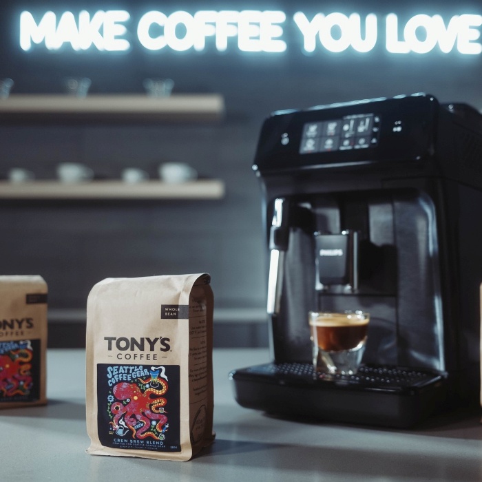Make Coffee You Love white Custom Neon® slogan sign on a black wall with a bag of coffee beans and a coffee machine in the foreground @seattlecoffeegear