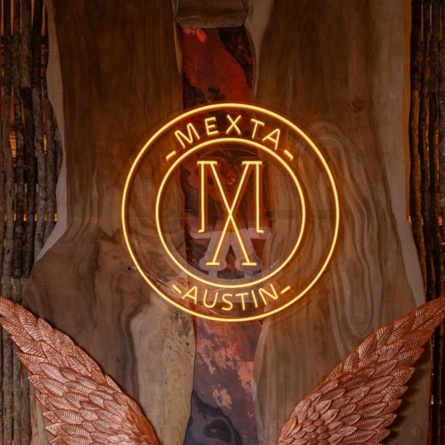 Warm white Custom Neon® 'Mexta Austin' circular logo sign on a live-edge wood wall above copper wing art