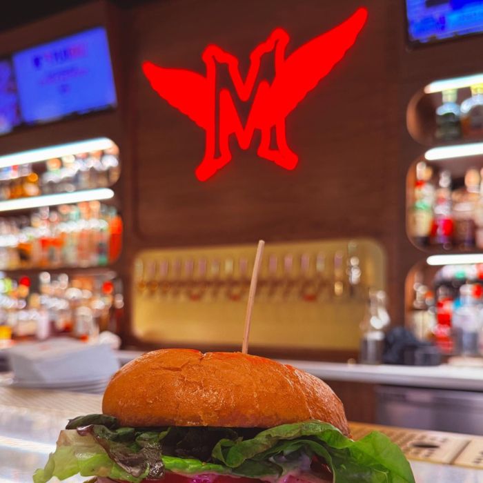 Burger on a bar counter beneath a glowing red custom neon winged 'M' logo, with beer taps and a stocked bar behind
