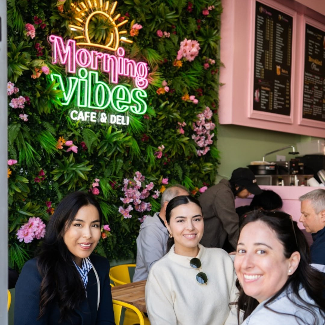 Morning Vibes Cafe and Deli colorful Custom Neon® logo sign on a lush plant wall with diners in the foreground