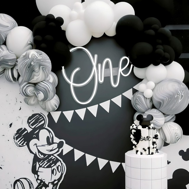 Custom Neon® One milestone birthday sign on a black background surrounded by a black and white balloon arch with a Mickey Mouse themed cake in the foreground - monochrome event decor by @decorandfiesta