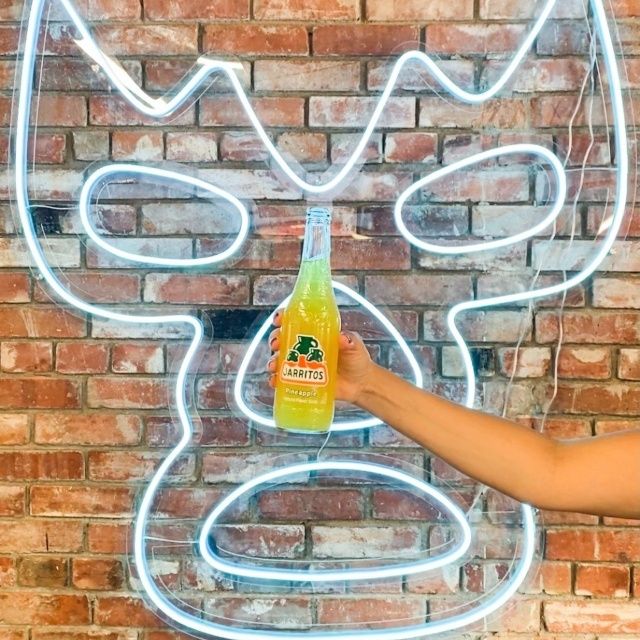 Hand holding a Jarritos pineapple soda in front of a white Custom Neon® luchador mask sign on a red brick wall