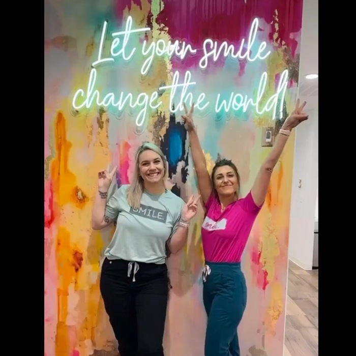 People posing in front of a Custom Neon 'Let your smile change the world!' sign on a multicolor painted wall