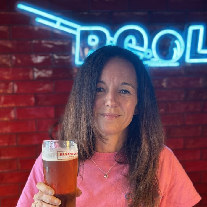Woman in a pink Custom Neon t-shirt holding a pint of Davenports lager in front of a blue custom LED neon 'Pool' sign on a red brick wall