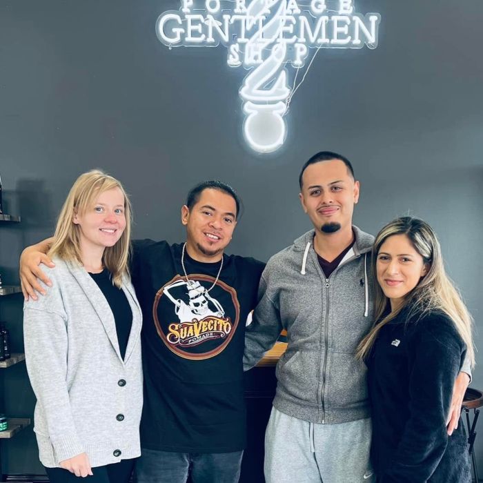 Four people posing in front of a white Custom Neon® sign on a grey wall that reads Portage Gentlemen Shop with a barber pole and top hat
