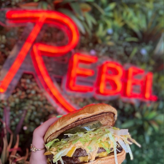 REBEL red Custom Neon® restaurant name sign shown on a lush greenery wall with a burger @rebelkitchenbar