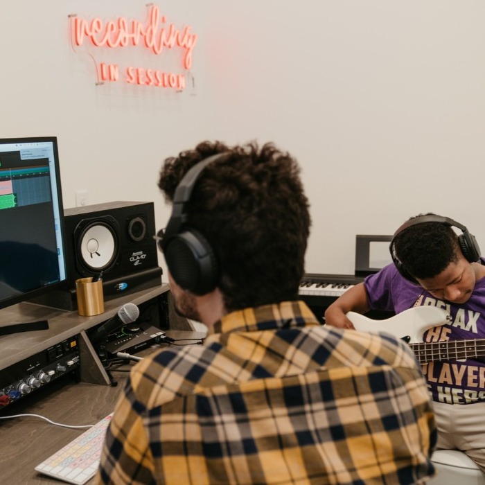 Red custom neon 'Recording in Session' sign on the wall of a studio where a man mixes at a desk, and a boy plays bass guitar